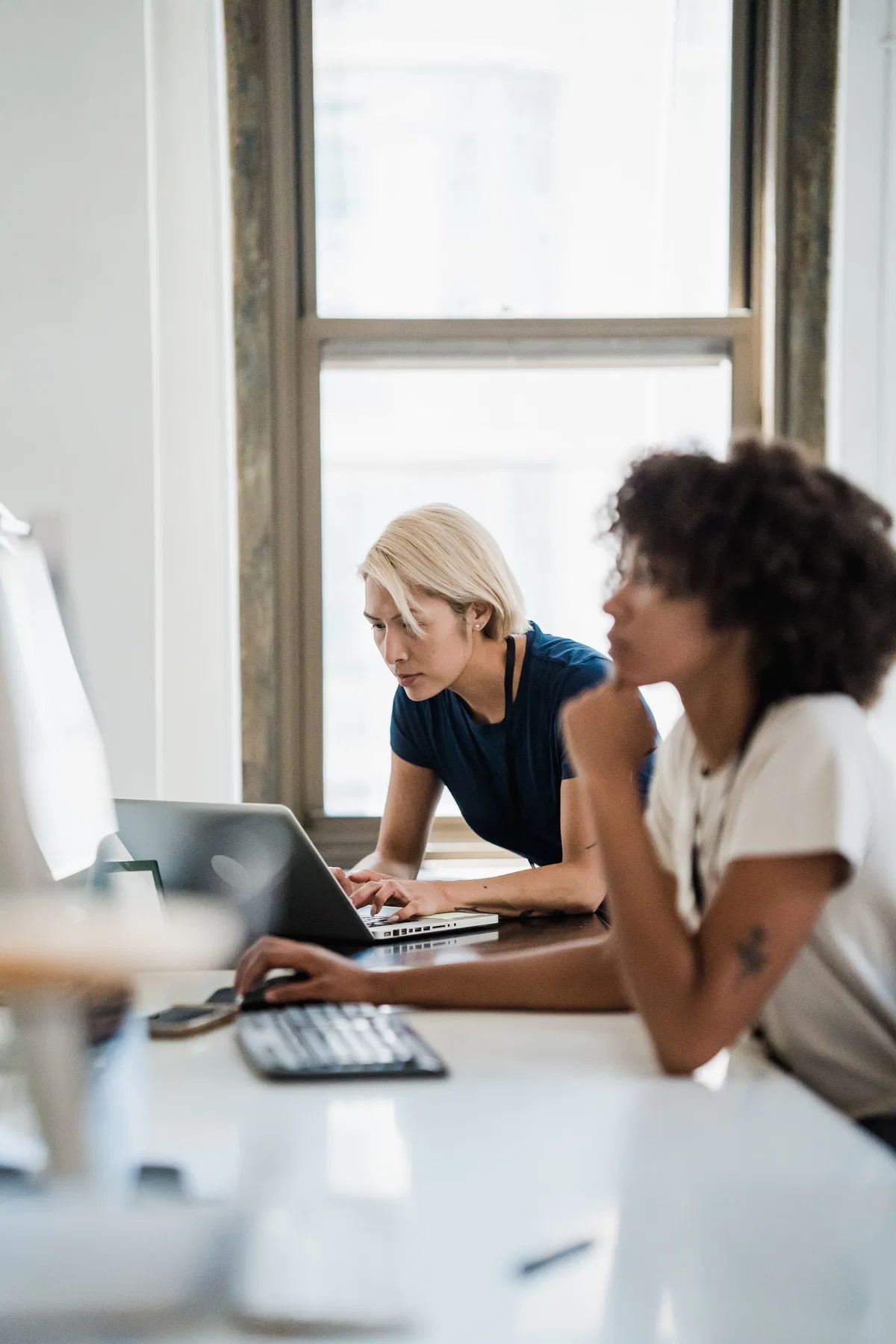 Two colleagues work at a long desk by a tall window, one leaning over a laptop.