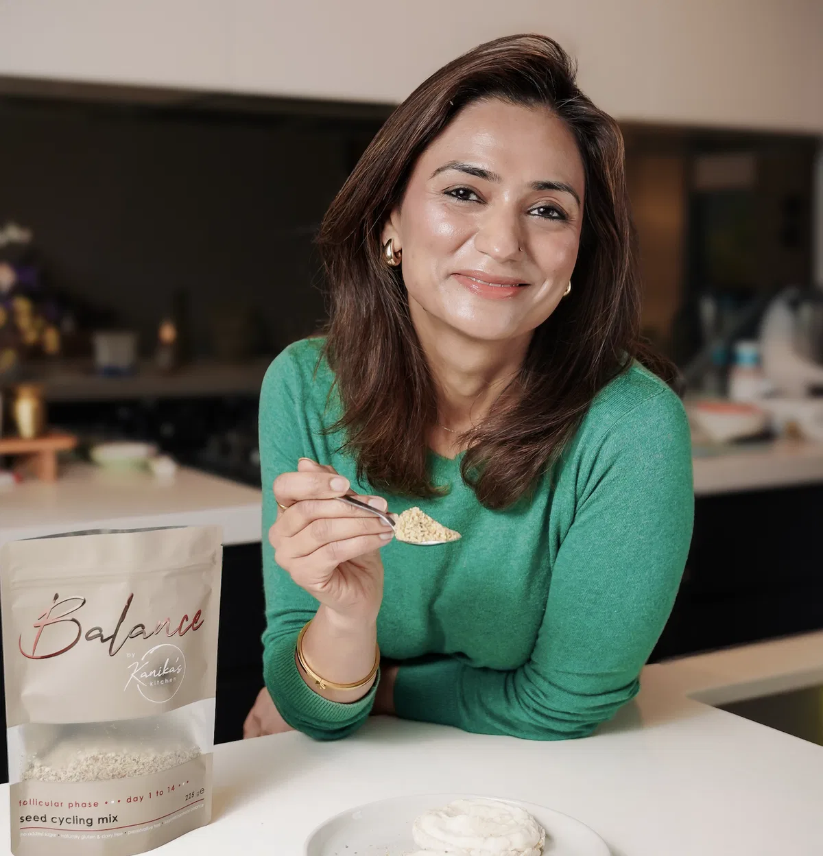 A founder smiles at her kitchen counter, a spoonful of her seed-cycling mix in hand and a bag of it beside her.