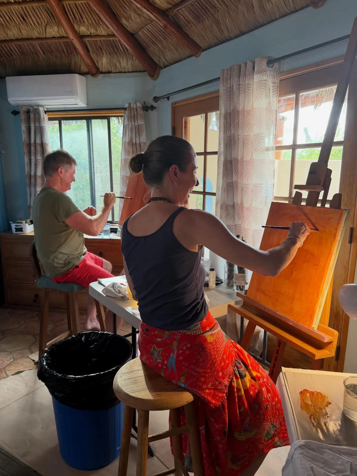 Two people paint at easels in a sunlit studio under a thatched roof.